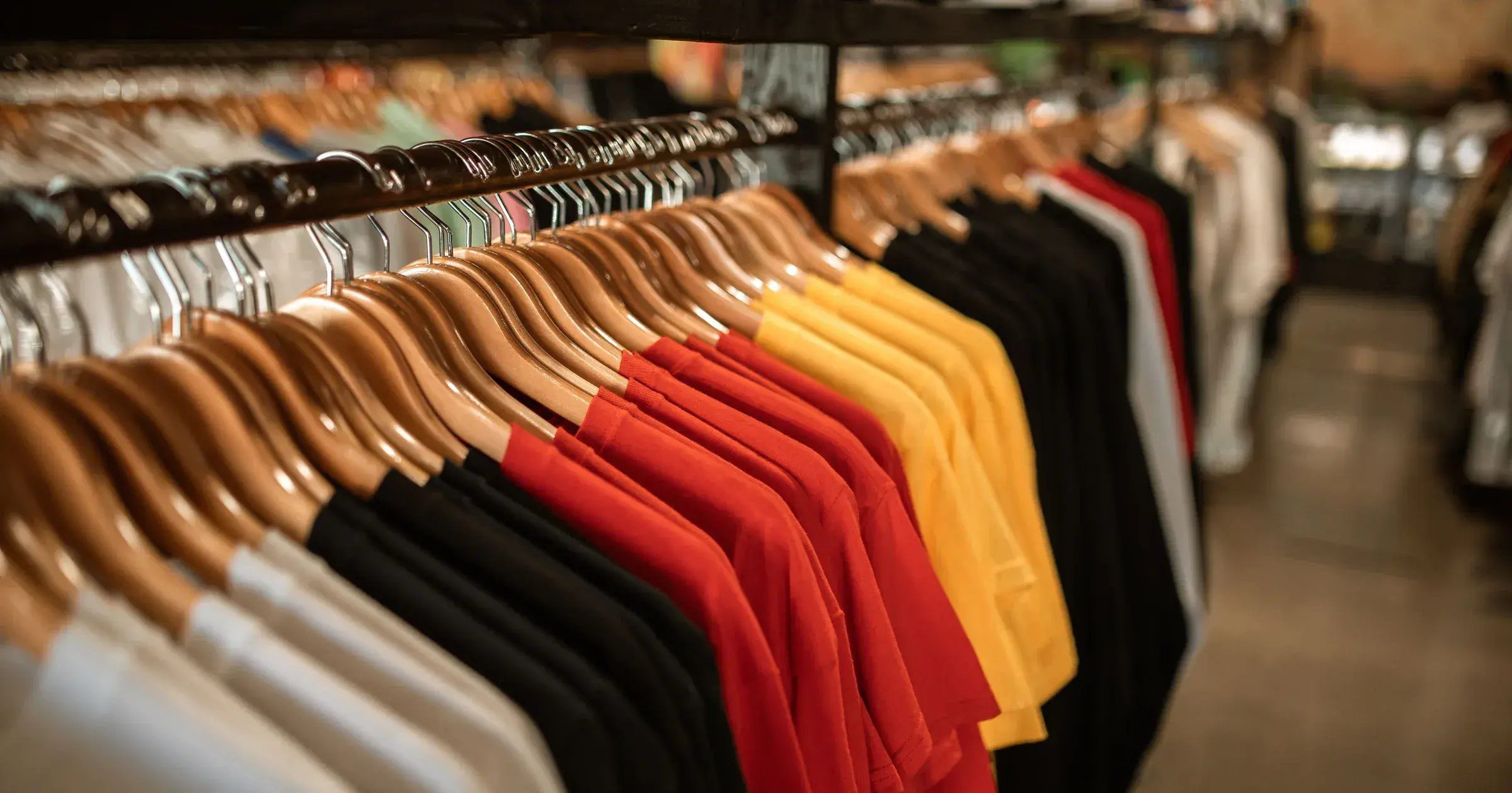 Rows of plain t-shirts and polos in white, black, red and yellow hanging on wooden hangers along a garment rail, illustrating how many colorways an apparel assortment carries