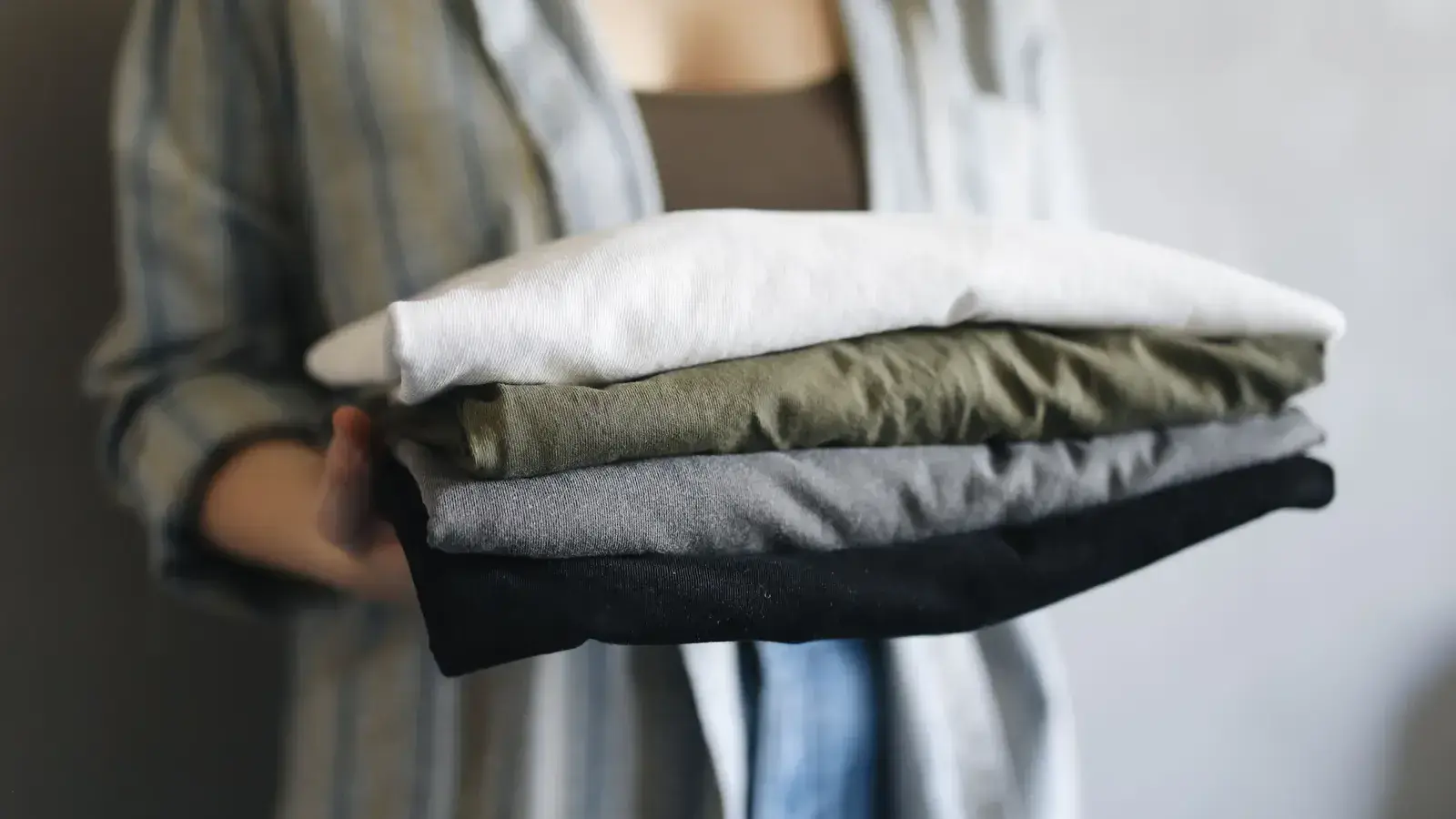 Person holding four folded shirts in neutral colors
