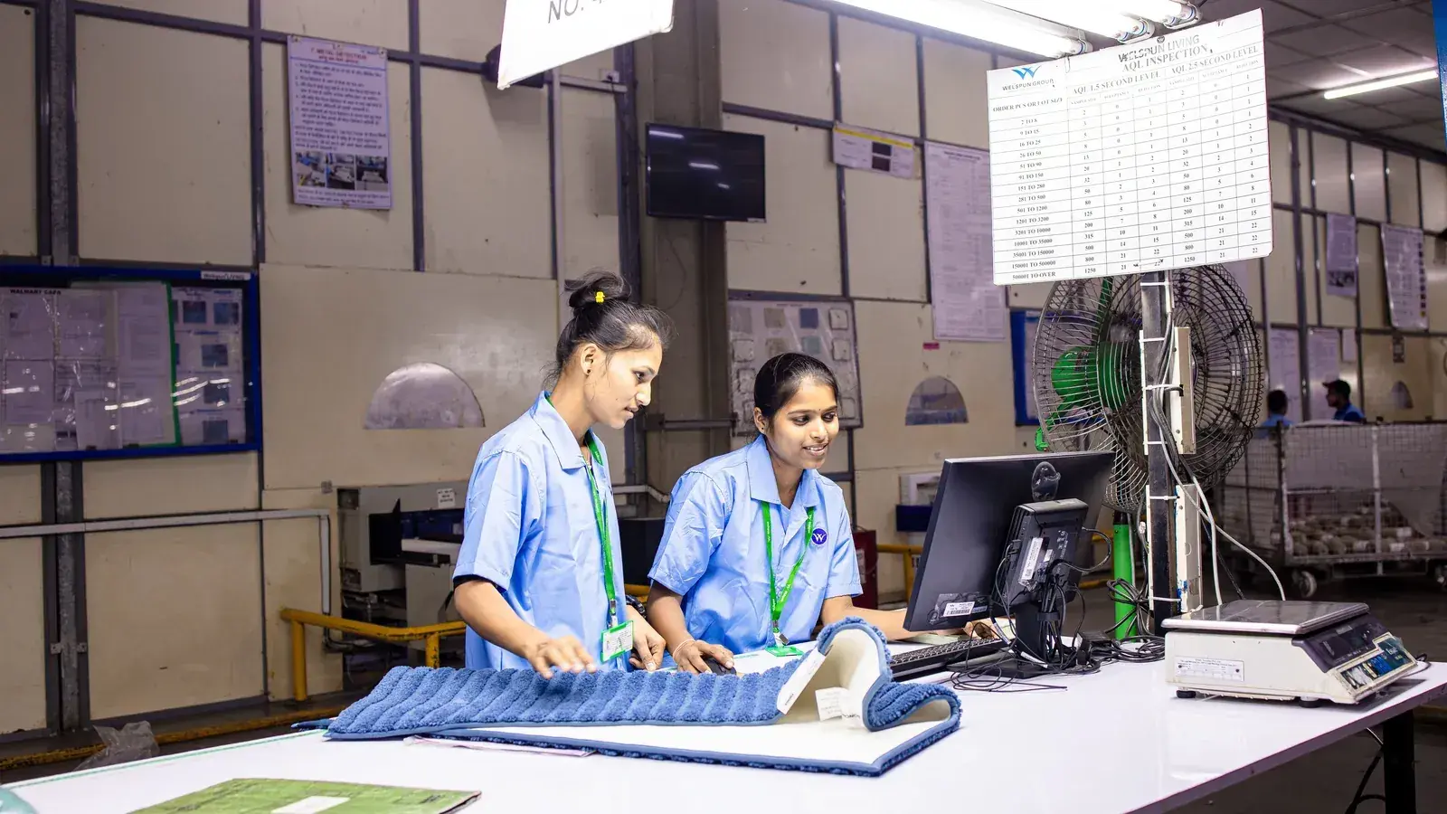 Two quality controllers working at an AQL inspection station with classification charts on the wall