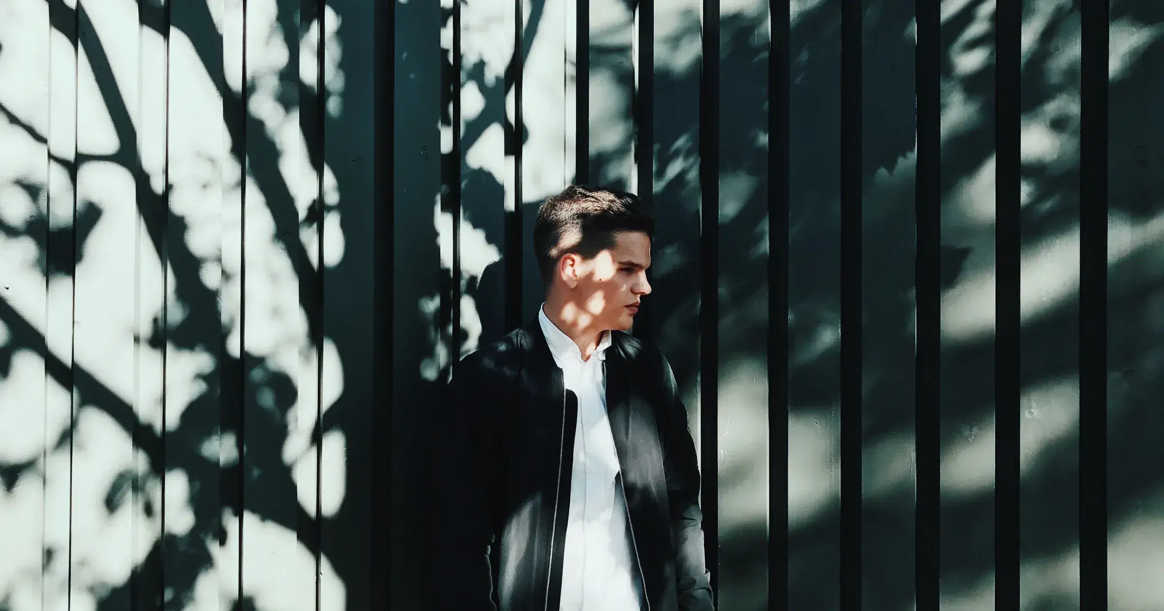 Man in an open black bomber jacket standing against a shadowed metal fence