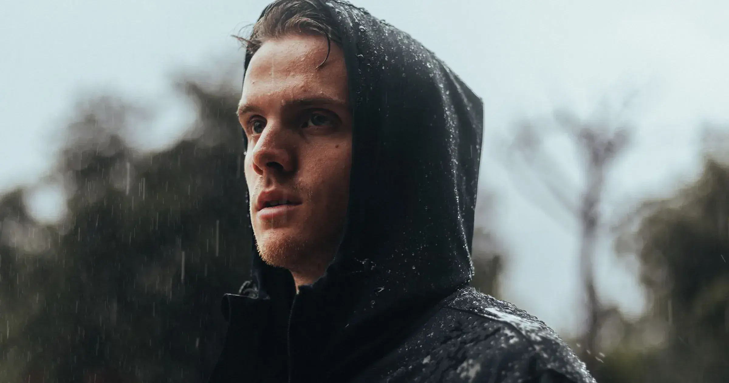 Man in a hooded black rain jacket standing in rainfall outdoors