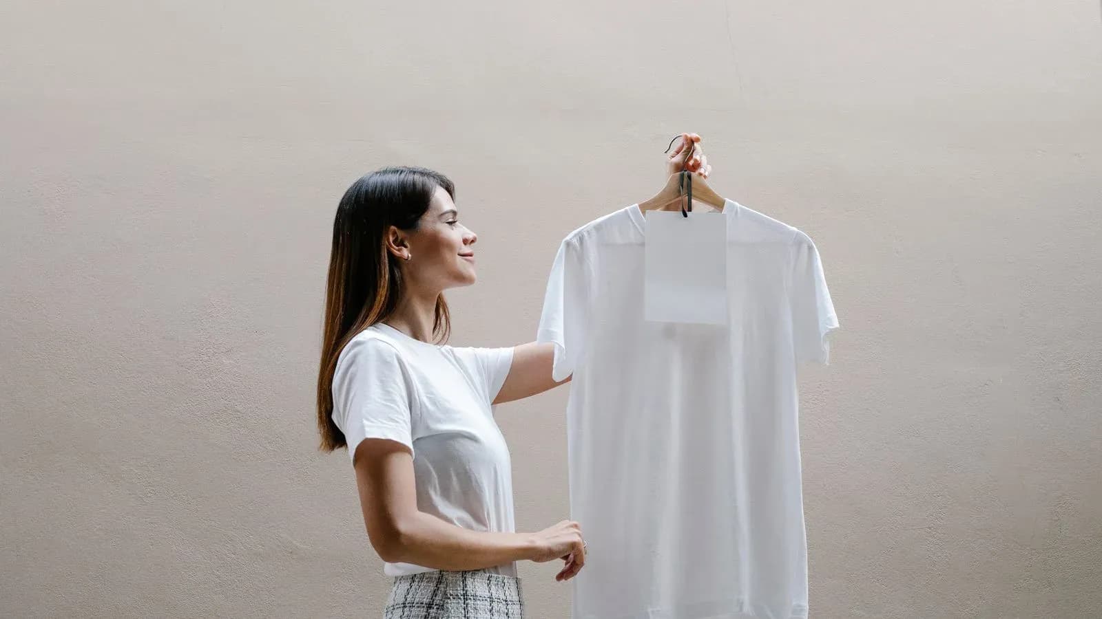 Woman holding a plain white T-shirt on a hanger against a neutral wall