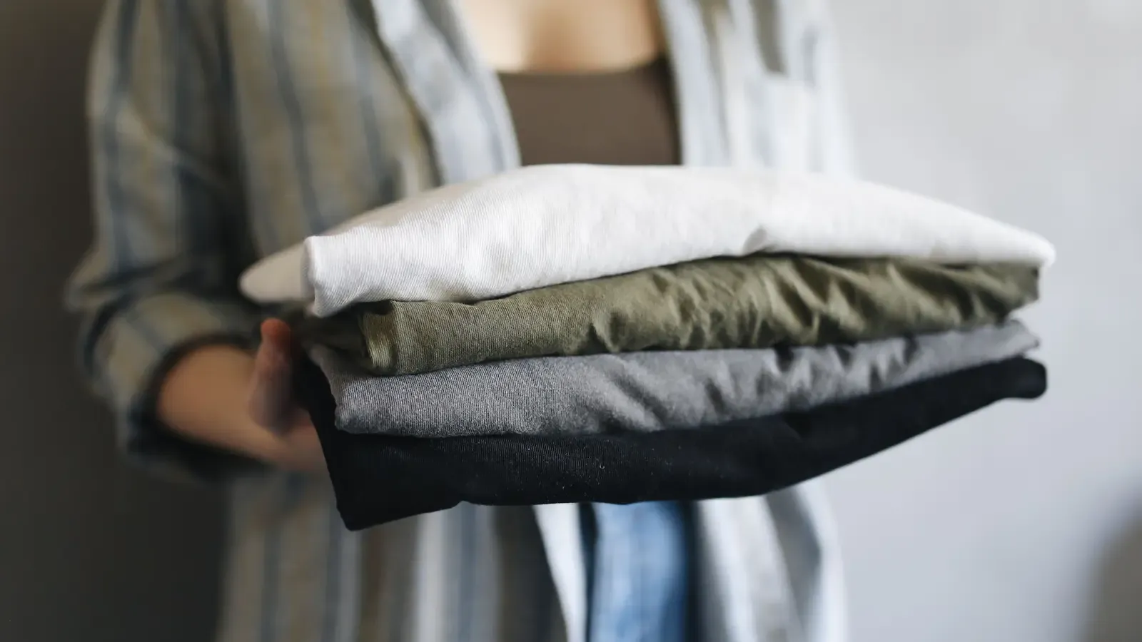 Person holding four folded shirts in neutral colors