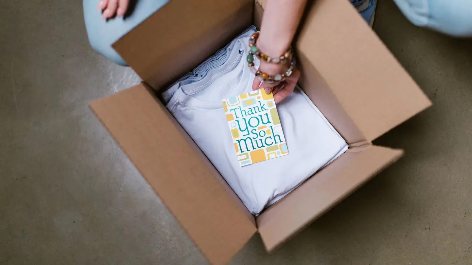 Person placing a folded white T-shirt and thank-you card into a cardboard box