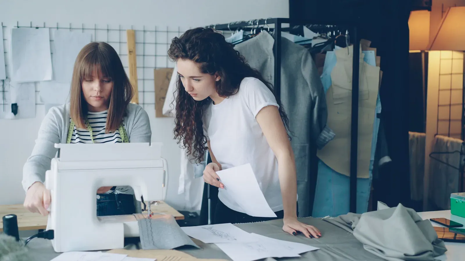 Two people working on a sewing sample in a design studio, with sketches and hanging garments nearby