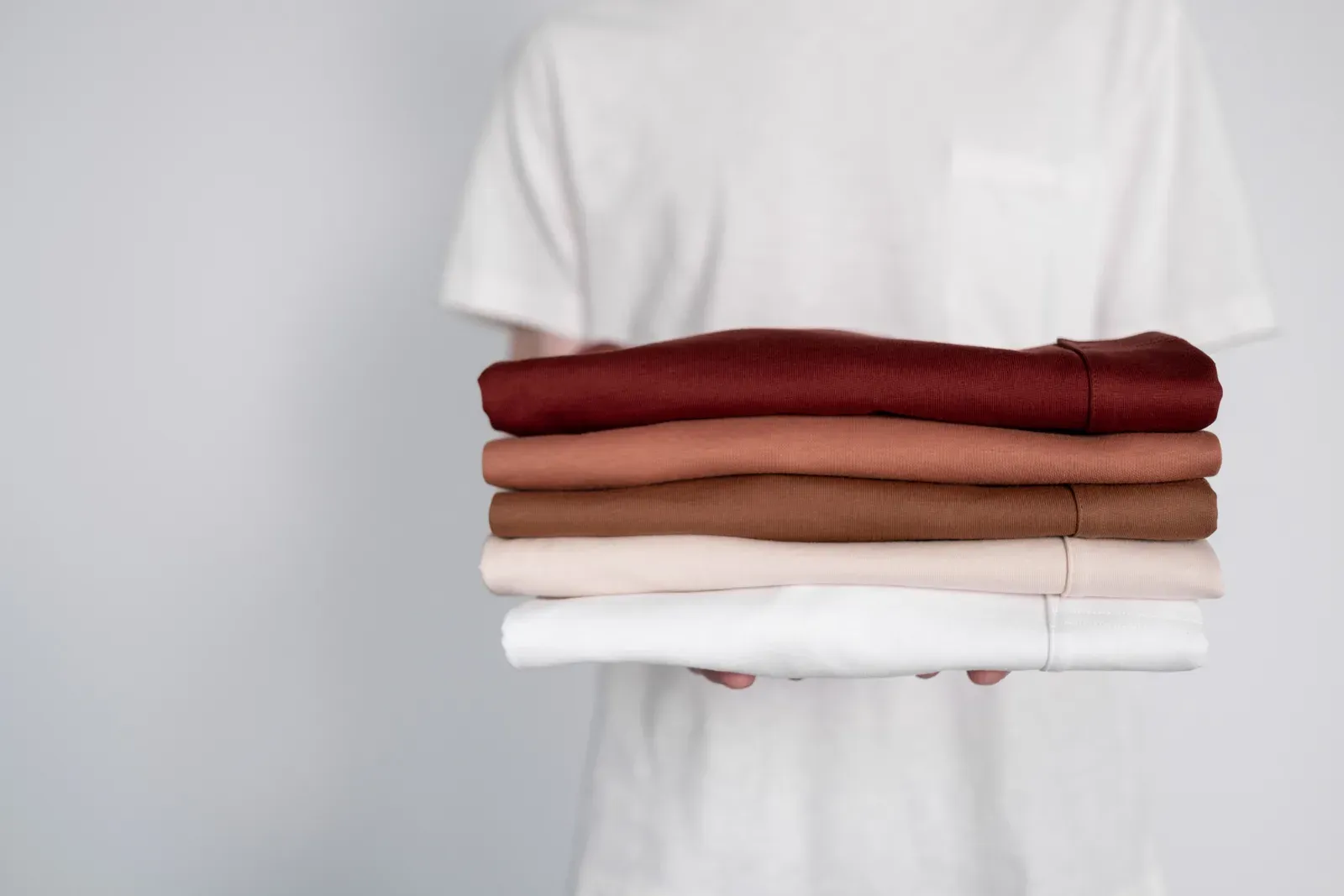 A person holding a neat stack of folded shirts in earthy colors
