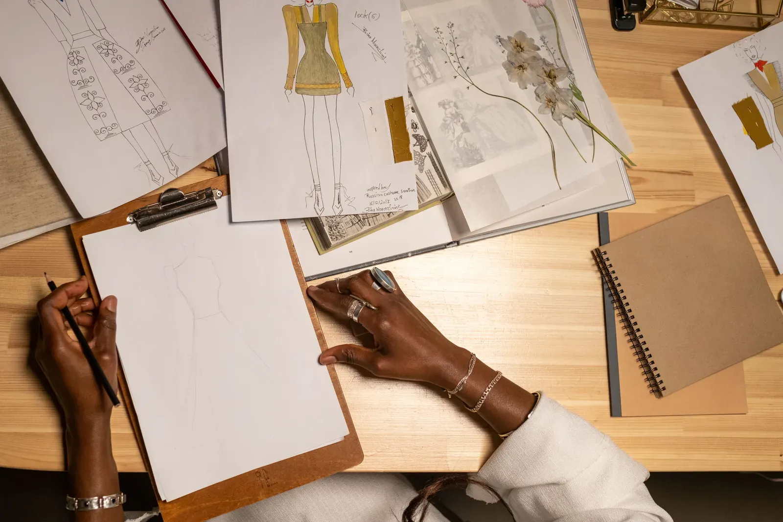 Overhead view of hands sketching a fashion design on paper, surrounded by reference sketches and fabric swatches