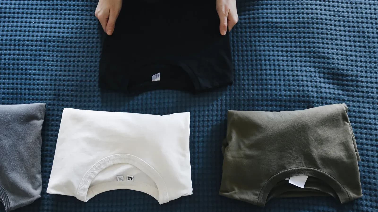 Four folded t-shirts in black, grey, white and olive laid out in a row while a pair of hands places one down