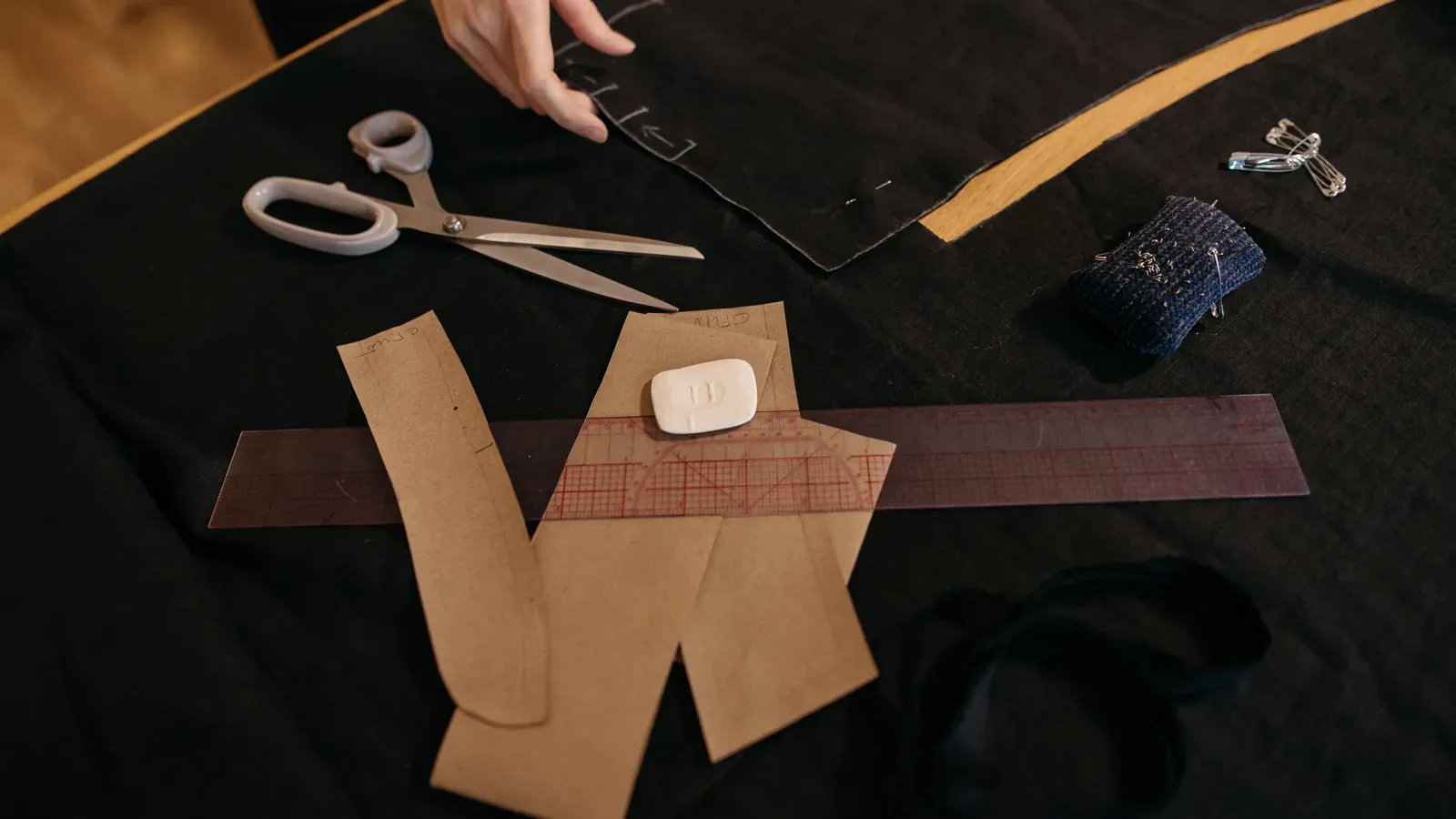 A paper pattern piece, scissors, a ruler and tailor's chalk laid out on a dark worktable