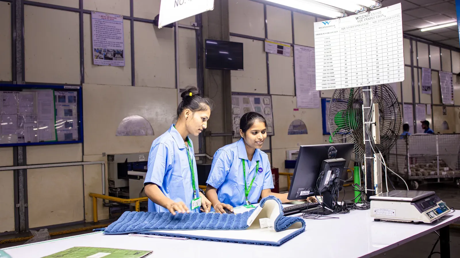 Two quality controllers working at an AQL inspection station with classification charts on the wall