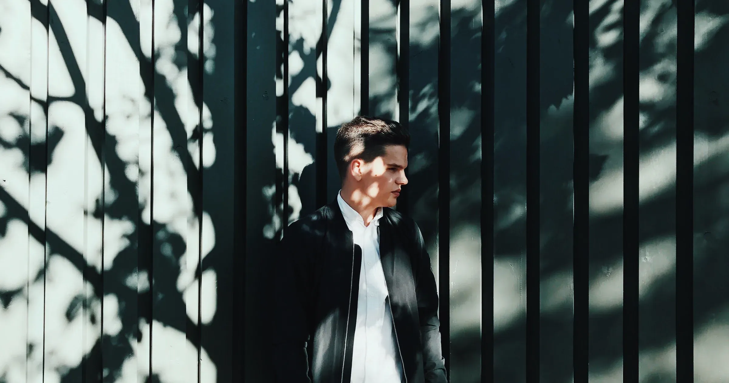 Man in an open black bomber jacket standing against a shadowed metal fence