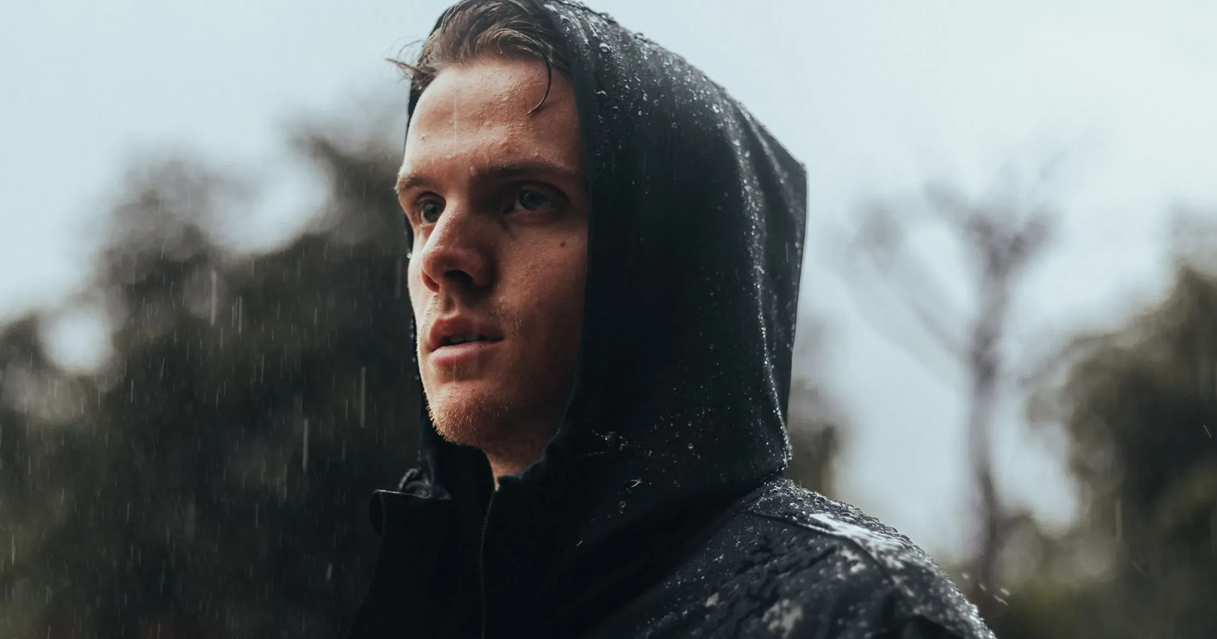 Man in a hooded black rain jacket standing in rainfall outdoors