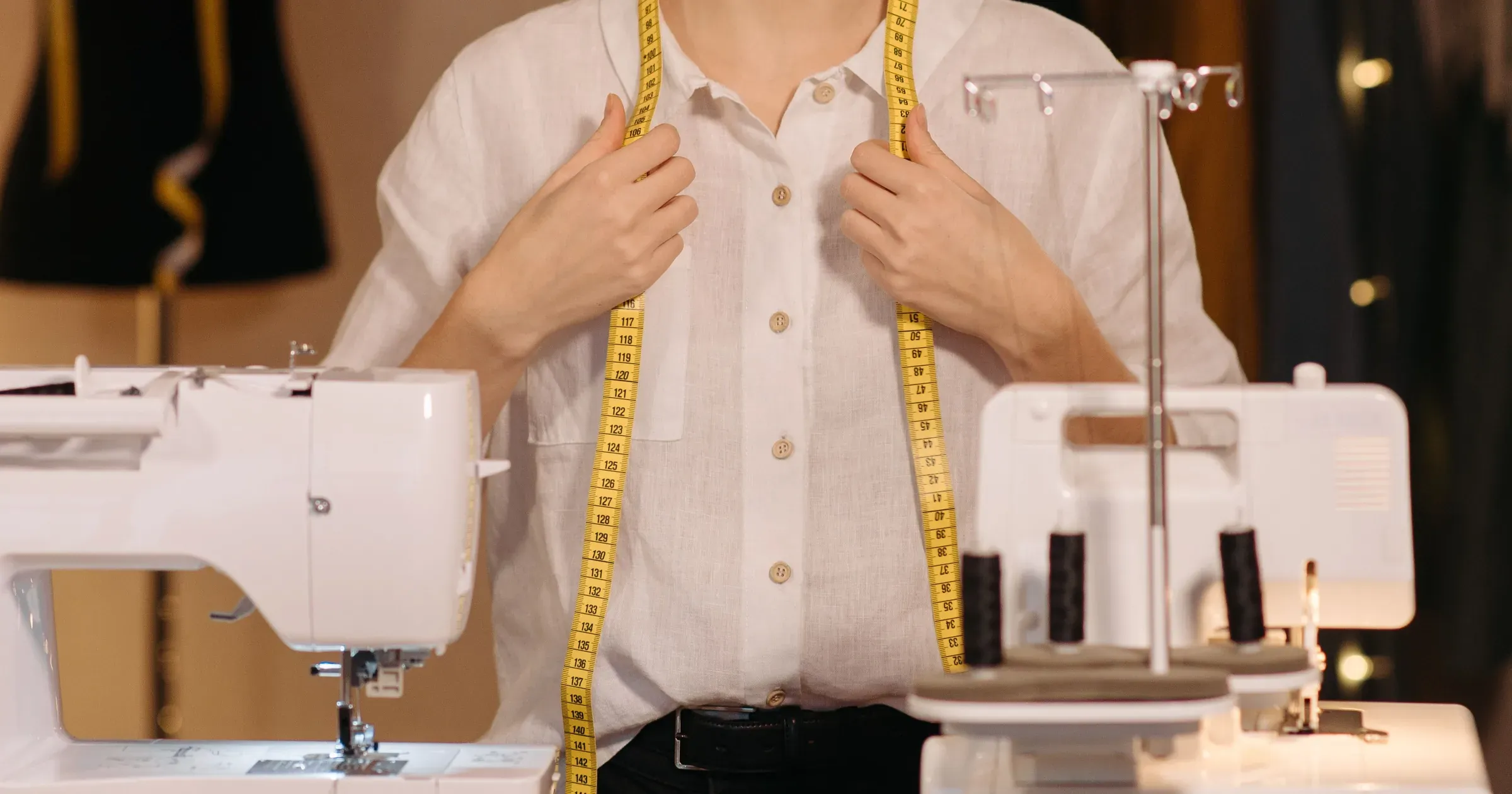 Garment worker in a white shirt holding a tape measure between two sewing machines in a workshop